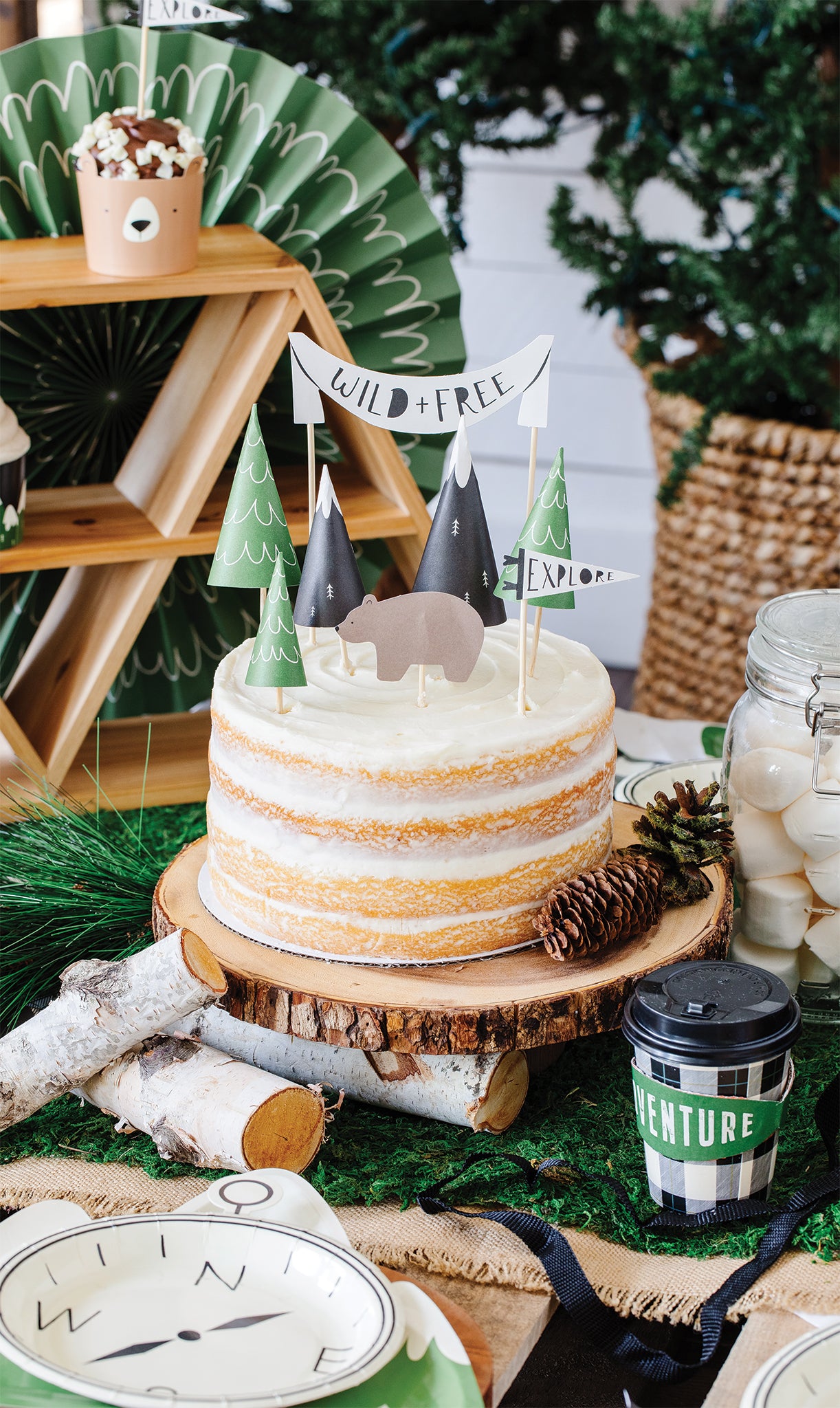 8 individual adventure./ happy camper cake topper. 3 x green mountain trees, 2 black and white mountain tops, 1 x Wild and Free mini cake topper banner, 1 x bear, 1 x explore pennant flag placed on a birthday cake.